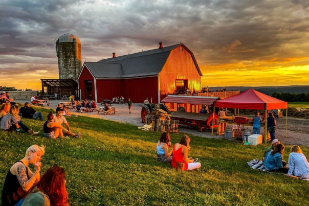 a sunset in back of a red barn with people sitting on the lawn in the foreground