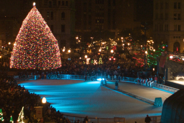 A bright lit up Christmas tree at night time overlooking an empty ice skating rink surrounded by a mass of people.
