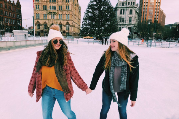two young women ice skating infront of historic city buildings and Christmas tree while holding hands