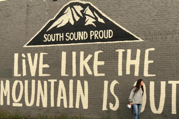 A person stands beside a Sumner mural reading “South Sound Proud” and “Live Like the Mountain Is Out.”