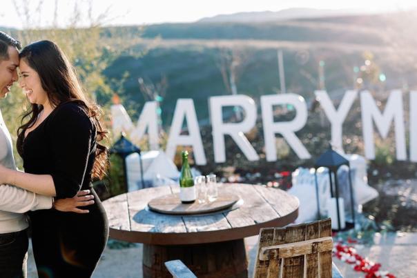 A couple embracing outdoors with a sign that read's "Marry Me"