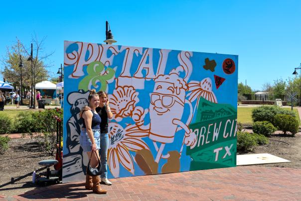 Two girls standing in front of a  temporary art mural depicting an artist's rendition of how they picture Temple being Brew City at Petals and Pints event in 2026.