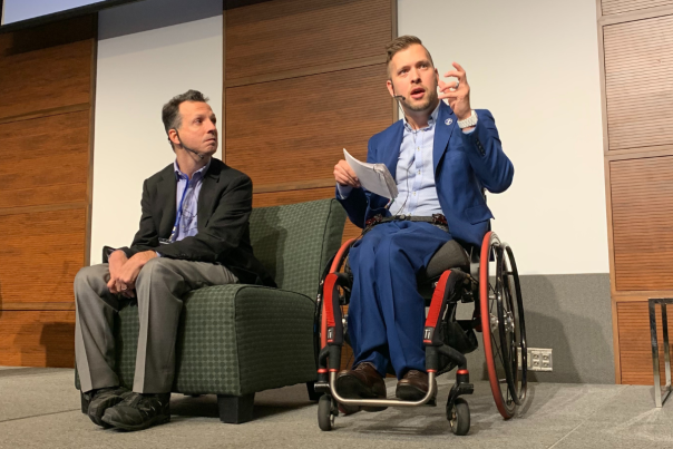 Two men are on stage, one holding notes and speaking to the audience from a wheelchair, the other man listening from a chair.