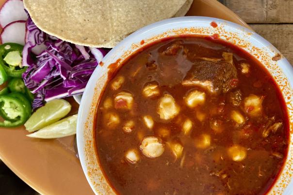 Close-up of a bowl of crimson red pozole rojo with hominy and tender pork in rich broth, served with a side of lime wedges, sliced radishes, shredded purple cabbage, jalapeños, and tostadas at Alta Baja Market in Santa Ana, California.