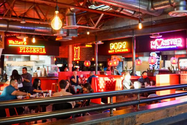 Interior of a retro, food court restaurant with people sitting at tables under bright neon restaurant signs