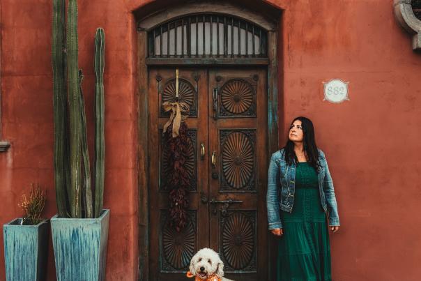 Woman with a dog standing in front of an orange-red Spanish style home with an ornanted wood door and potted cacti