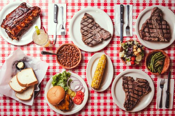 top view of a table with plates of steak, ribs, corn on the cob, beans, burgers, bread, and butter