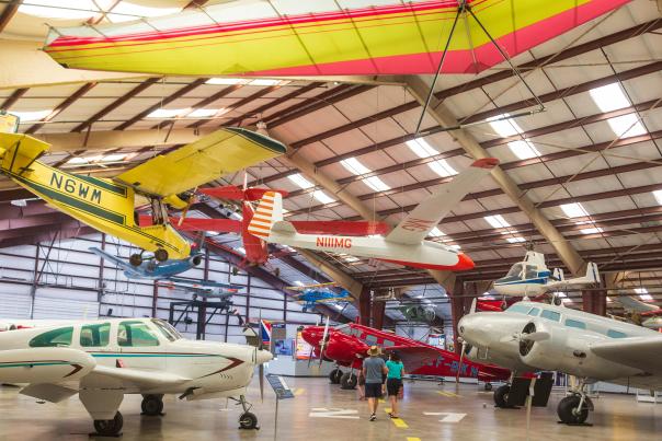 two people walking in the interior of a plane hanger with several different types of planes on the ground and suspended from the air