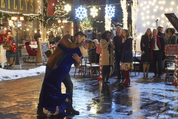 A festive street scene from a holiday movie, featuring a man dipping a woman in a romantic embrace in the center of a charming, snow-dusted setting. The woman is wearing an elegant blue gown, while the man is dressed in a warm brown jacket. Around them, twinkling lights, decorated storefronts, and giant hanging snowflakes create a magical winter atmosphere. A small crowd of onlookers dressed in festive attire smiles and claps in the background, adding to the celebratory mood.