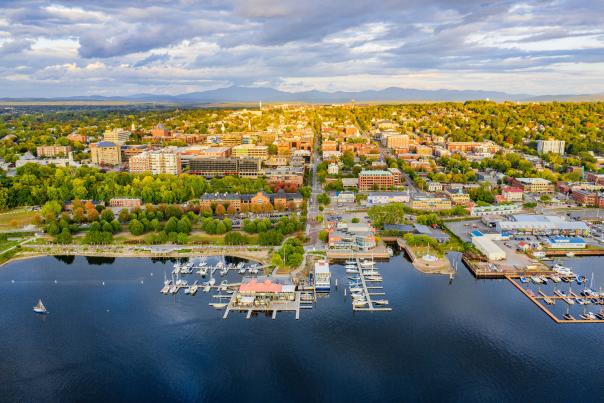 Aerial view of Burlington by a lakeshore with marinas, docks, and numerous buildings. Greenery surrounds the area, and mountains are visible in the background.