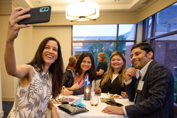Three women and one man capturing a selfie at an event gathered around a table of beverages and appetizers.
