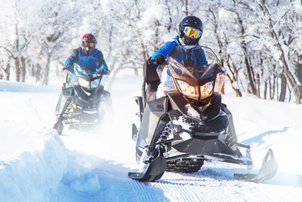 two people snowmobiling on snowy trail with trees in background