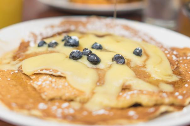 A close-up of a plate of pancakes topped with blueberries and syrup, with a blurred figure in the background pouring syrup. Glasses of orange juice are visible.
