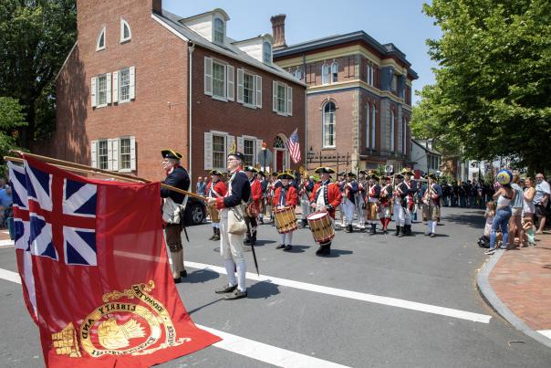Revolutionary War reenactors, some carrying a large red British flag, march down a street lined with historic brick buildings and spectators on a sunny day.