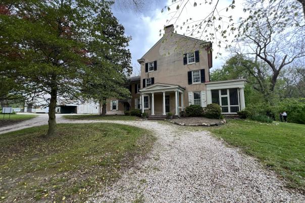 Two-story historic house with a portico entrance, set back from a gravel driveway, with a white barn visible to the left and trees around.