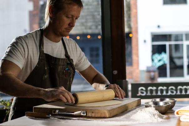 blonde male chef rolling dough on wood board with wooden rolling pin, wearing dark brown apron