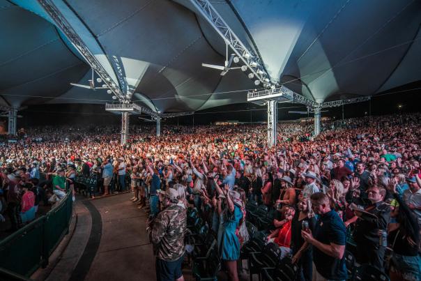 A wide-angle shot of a full-house audience at The Cynthia Woods Mitchell Pavilion. Many attendees are wearing Western clothes (denim clothes, cowboy hats, and boots). Spotlights sway over the crowd.