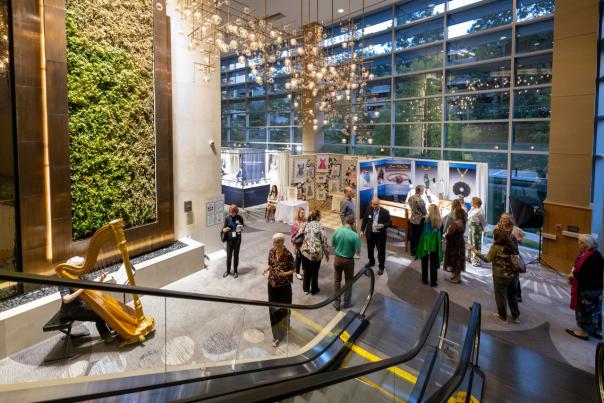 The camera overlooks a fancy hotel lobby from the escalator. On the left, an experienced woman plays a golden harp in front of a wall of decorative moss. Arcing across the image to the right are booths displaying vendors' fine craft pieces. Potential buyers stand around, gazing at the art or listening to the harp music.