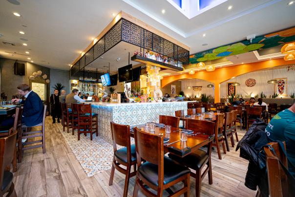 The camera reveals a wide-angle shot of the Xalisko dining room and bar from the front left corner of the building. There are a number of solid wooden tables that seat and are plated for four. The bar is covered in white, brown, and turquoise tiles shaped like flowers. In the back right of the frame is the rest of the dining room. The back wall features images of colorful corn cobs. On the ceiling is a long art piece of many interconnecting crocheted hexagonal panels. The panels alternate in colors of turquoise, lime green, dark forest green, and orange.