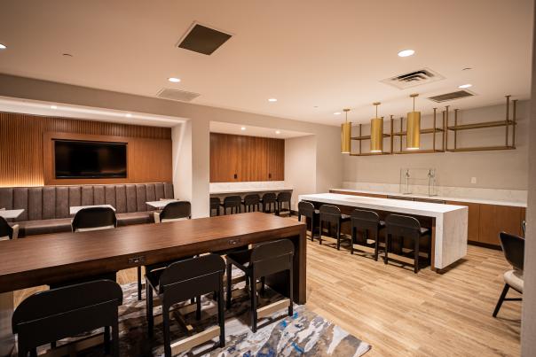 The camera overlooks a spacious, clean, modern-looking room with ample counter space along two walls, plus two-person booths along two more walls and two long tables positioned perpendicular from each other in the middle of the room. The table along the far wall seats four, and the one in the middle seats six.
