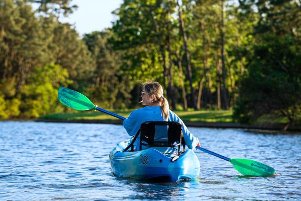 A young blonde woman (Brooklyn Newell) holds a black and green double-sided paddle to row her bright blue kayak. She is facing away from the camera, paddling along Lake Woodlands toward a copes of trees.