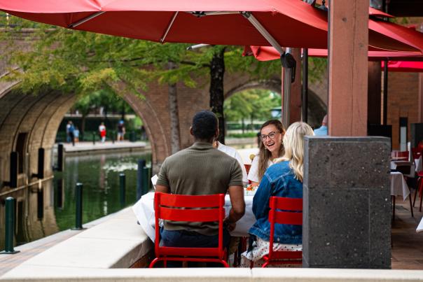 Four young adults (two with their backs to the camera) sit at a patio table with red metal chairs and a large red square umbrella hanging overhead. The table is on an elevated patio overlooking The Waterway. In the background is the Waterway Avenue bridge.