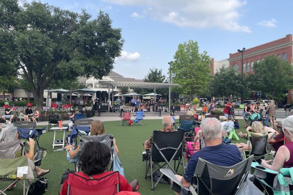 The camera faces the stage from the back of Market Street's Central Park during National Travel and Tourism Week 2024. Between the camera and the stage are a bunch of spectators sitting in the grass, in lawn chairs, or under white pop-up tents.