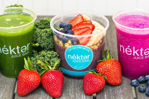 The camera faces a wooden picnic table featuring three plastic cups bearing the "nékter JUICE BAR" label. The cup on the left is filled with a bright green smoothie and surrounded by broccoli stalks. The cup in the middle contains a dark purple-ish smoothie topped with oats, blueberries, strawberries, and banana slices. The cup on the right is full of a bright magenta (dragonfruit?) smoothie surrounded by strawberries and blueberries.