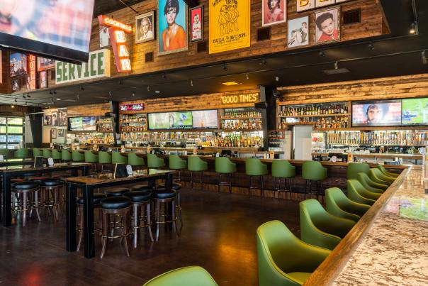 The image depicts the interior of Kirby Ice House, featuring a row of wood tables with six round bar stools each and a LONG bar with at least 20 green bar chairs. The walls are decorated with neon signs, vintage photos of celebrities, and other memorabilia.