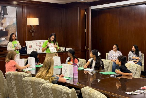 Noemi Gonzalez teaches a workshop during the 2026 Young Makers Market. A diverse group of 6-8 students (approx. 8-10 years old) listens intently. The workshop is set in a fancy conference room with dark wood walls and plush chairs. Some of the children's parents observe from the doorway.