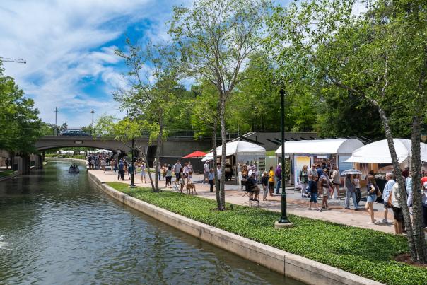 White tents and umbrellas line The Woodlands Waterway to shade displays of local and international artwork for sale at The Woodlands Waterway Arts Festival. Dozens of people of all ages take in the sights.