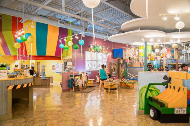 A wide-angle view of the interior of The Woodlands Children's Museum. The walls are painted purple with thick, multi-colored stripes. Light displays look like clouds and paper lanterns. There are exhibits with toy trains, a farmer's market, ships on the ocean, and more.