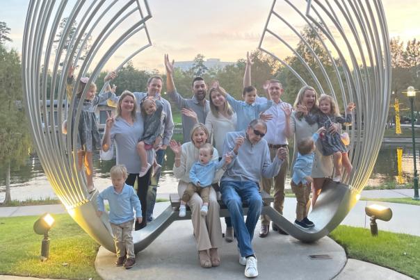 The camera faces the Memorial Hermman The Woodlands Medical Center's "Proud Souls" art bench, which looks like two large metal waves or wings spreading inward over the bench. Sitting on the bench is Linda Nelson and her husband. Behind and around them are their three children with their partners and seven children. Everyone is dressed in dressy-casual clothes in some shade of white, gray, or pale blue, and everyone (except a few runaway toddlers) is smiling and waving at the camera. In the background is The Woodlands Waterway and a beautiful wooded area on the opposite shore.