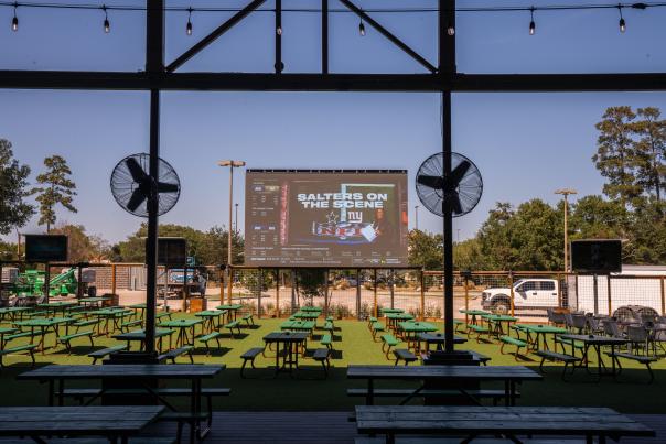 The Kirby Ice House Patio features dozens of green picnic tables, some shaded by the building, with large fans overhead and a giant projector screen showing sports stats in the back.