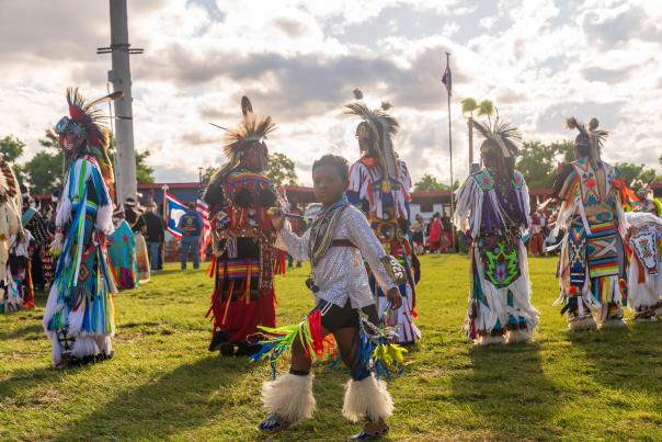 The scene is a sensory explosion of color and community, set against a grassy field under a brilliant, sun-drenched sky. In the background, the Wyoming state flag and the American flag fly, grounding this cultural event in its iconic regional landmark of Fremont County.