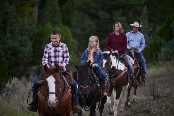 A family riding horses along a wooded trail, with two children in front and two adults following behind in an outdoor setting.