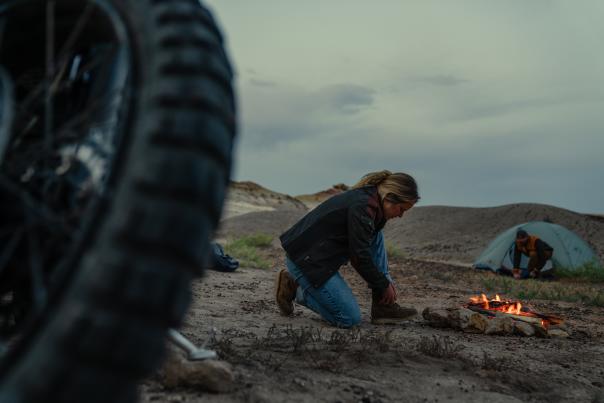 A person kneels by a small campfire in a desert landscape, with a motorcycle tire in the foreground and a tent nearby. The sky is overcast.