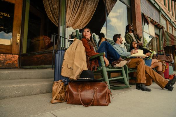 Four stylish travelers lounge in green rocking chairs outside the historic Occidental Hotel in Buffalo, Wyoming, surrounded by leather luggage and western attire during their travel across Wyoming.