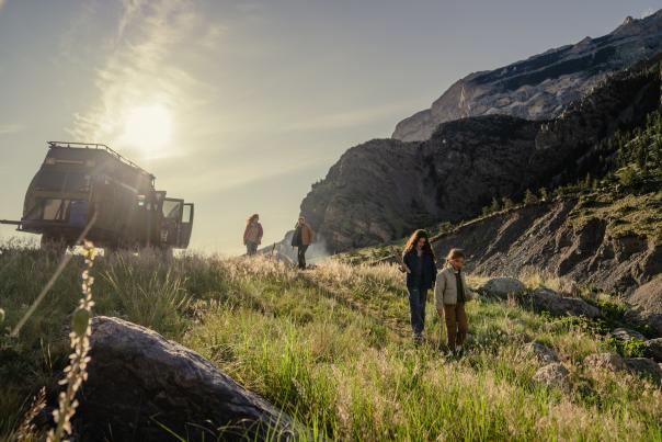 Four people walk down a grassy hillside near a parked off-road vehicle, with rugged mountains rising behind them in warm evening light.
