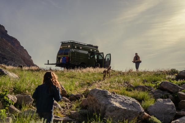 A green camper van is parked on a grassy hillside near rocky terrain under a bright sky. A man stands beside the open van doors while a dog runs toward him. In the foreground, another person with long hair walks uphill toward them, partially turned away from the camera.