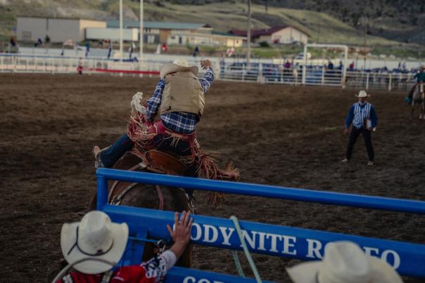 A rodeo rider in western attire leans forward while riding a bucking horse inside a dirt arena, holding on with one hand. Other cowboys in hats stand nearby behind a blue metal gate, watching and assisting, with buildings and hills visible in the background.