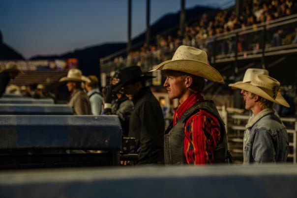 Rodeo cowboys in hats and western attire stand along the arena fence at dusk, with a crowd in the grandstands behind them, capturing Wyoming’s rodeo culture and community atmosphere.