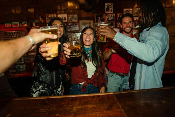 A group of friends gathers inside a rustic western bar, raising drinks in a celebratory toast. Warm lighting, wood-paneled walls, and framed photographs create a lively and welcoming atmosphere that reflects Wyoming’s social and nightlife culture.