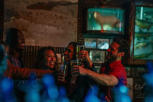 A group of four adults stands closely together inside a dimly lit bar, smiling and raising glasses in a celebratory toast. Warm, colorful lighting highlights their faces, while rustic decor and glass display cases with taxidermy animals are visible in the background, creating a lively and social Western nightlife atmosphere.