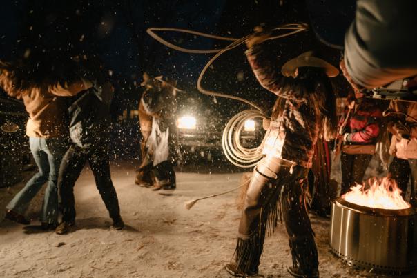 A cowgirl swings a lasso beside a glowing campfire during a snowy winter evening near Jackson, Wyoming. Surrounded by people in Western attire and illuminated by vehicle headlights, the scene highlights Wyoming’s cowboy culture, ranch traditions and winter gathering experiences.