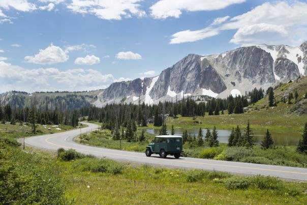 A green vehicle drives along a winding mountain road past a small lake, with pine trees and snow-dusted peaks rising under a bright blue sky.