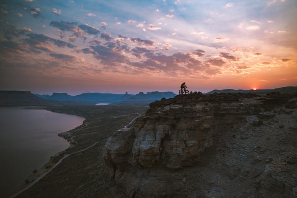 A cyclist rides along the edge of a rocky cliff at sunset, overlooking a wide river and canyon landscape under a colorful sky.