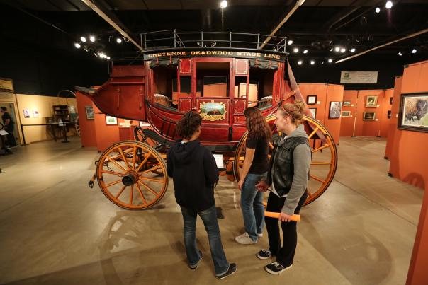Visitors explore the Cheyenne Frontier Days Old West Museum in Cheyenne, Wyoming, viewing the historic Cheyenne–Deadwood Stage Line coach on display. The exhibit showcases Wyoming’s transportation history and Old West heritage within a museum gallery featuring western art and historical artifacts.