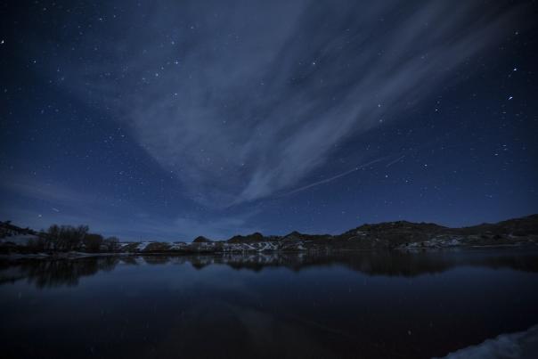 A star-filled night sky stretches above a calm lake, with wispy clouds and distant hills reflected in the still water.