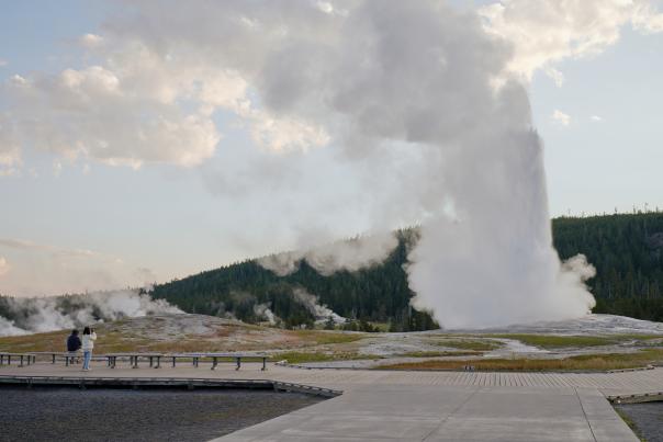 The Old Faithful geyser erupts with a tall column of steam in Yellowstone National Park as visitors watch from a wooden boardwalk.”